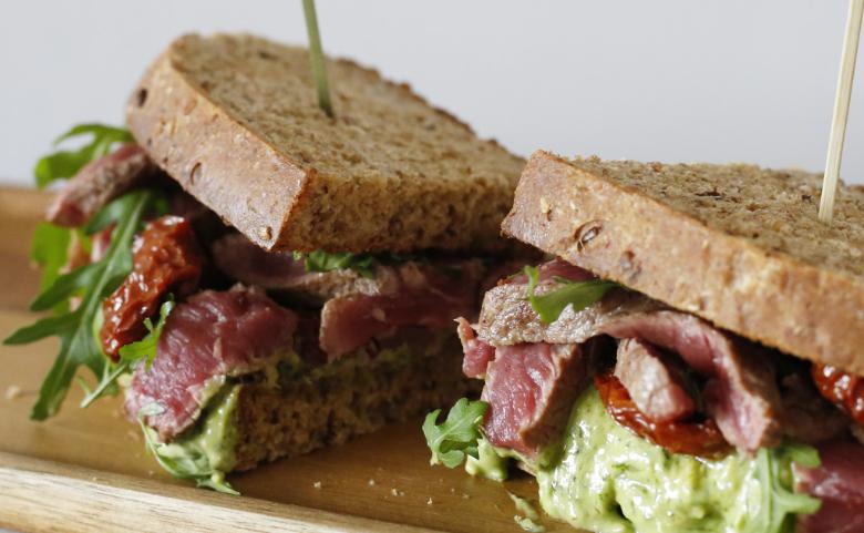Wholegrain bread sandwich filled with sliced rare steak, rocket, sun-dried tomatoes, and avocado spread